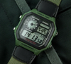 A Casio digital watch with an olive green and black rugged case and matching green-and-black strap, photographed close up on a dark textured fabric background.