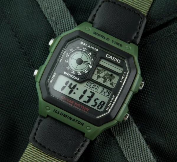 A Casio digital watch with an olive green and black rugged case and matching green-and-black strap, photographed close up on a dark textured fabric background.