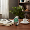 Cassims&Co. Casio Enticer watch with a green sunray dial and stainless steel bracelet on a desk with coffee and a book.