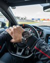 Cassims&Co. TAG Heuer Carrera Calibre 16 watch with brown dial on a driver's wrist inside a race car cockpit.
