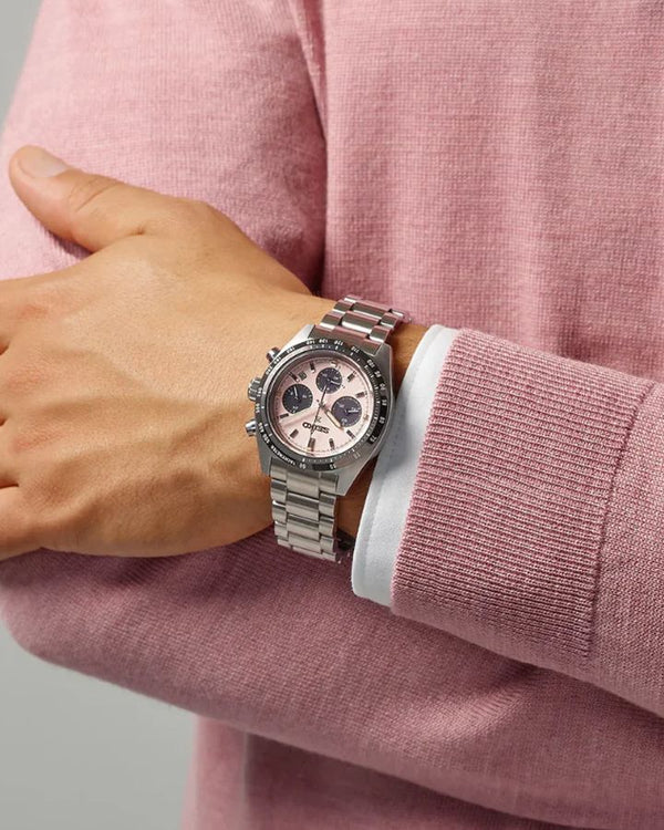 A silver stainless steel chronograph watch with a rose gold dial and matching rose gold bezel, worn on a person's wrist over a pink knit sweater.
Cassimsandco.